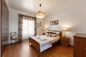 A hotel room with a wooden bed, two bedside tables with lamps, a chair, a wardrobe, and a window with curtains.