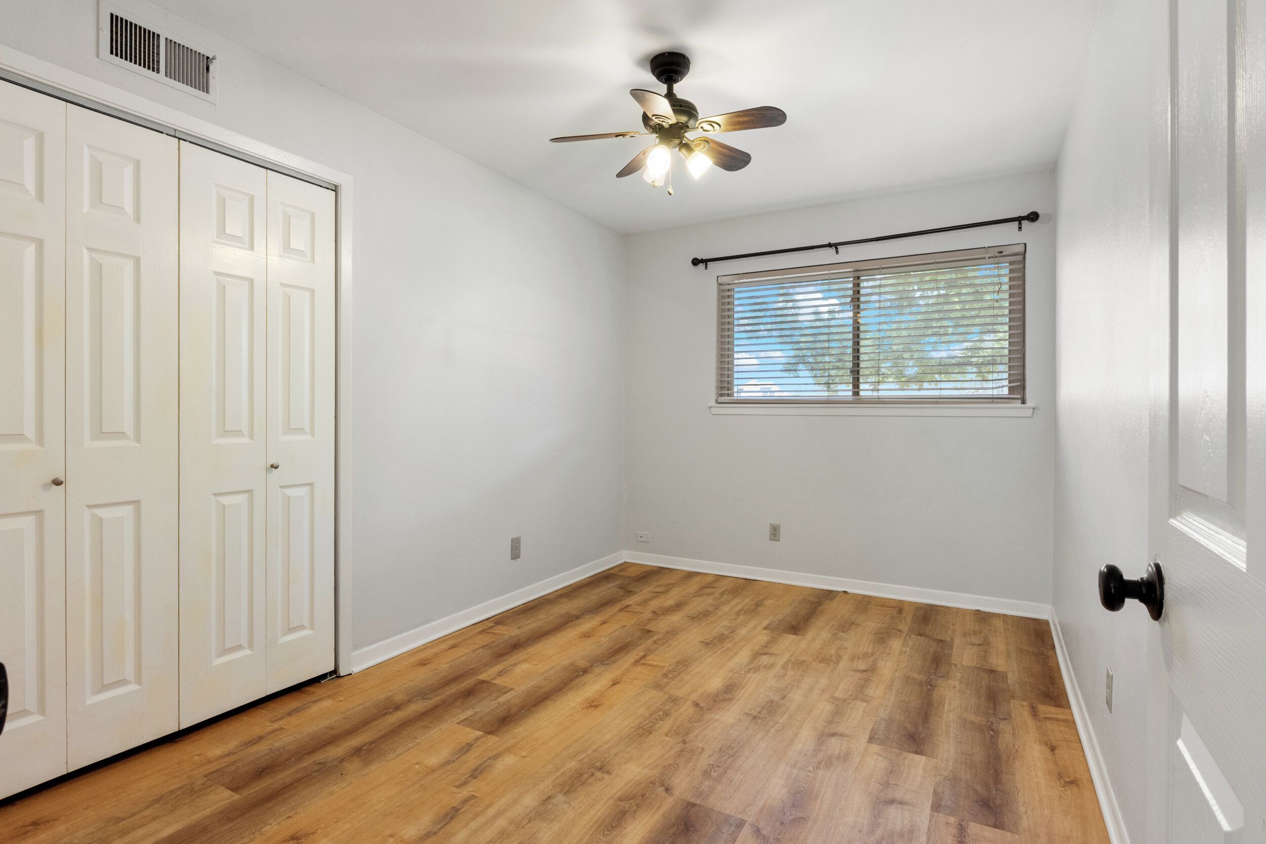 A room with white walls, a ceiling fan, a window with blinds, and a wooden floor.