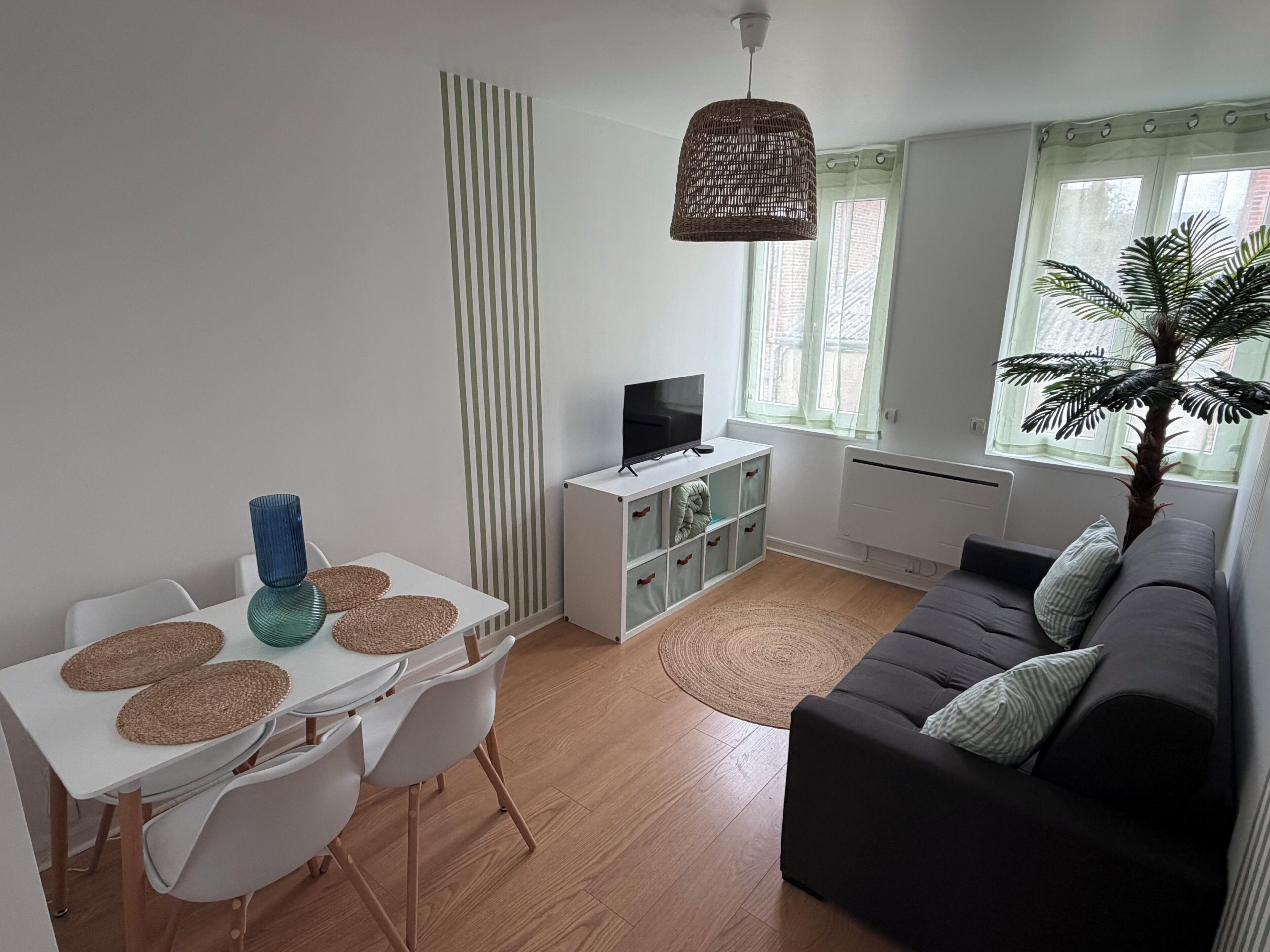 Ein Wohnzimmer mit einer Couch, einem Fernseher, einem Esstisch mit Stühlen, einer Pflanze und einem Fenster mit Vorhängen.