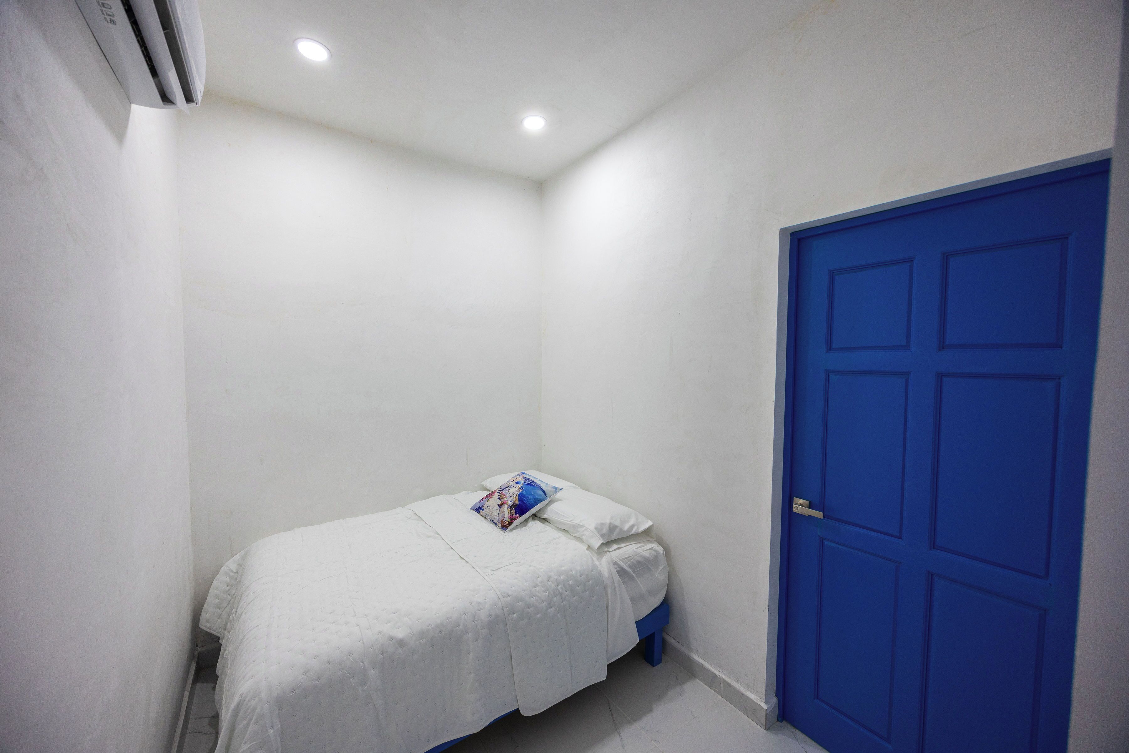 Une petite chambre avec un lit simple, du linge de lit blanc, une porte bleue et une bouche d’aération au plafond.