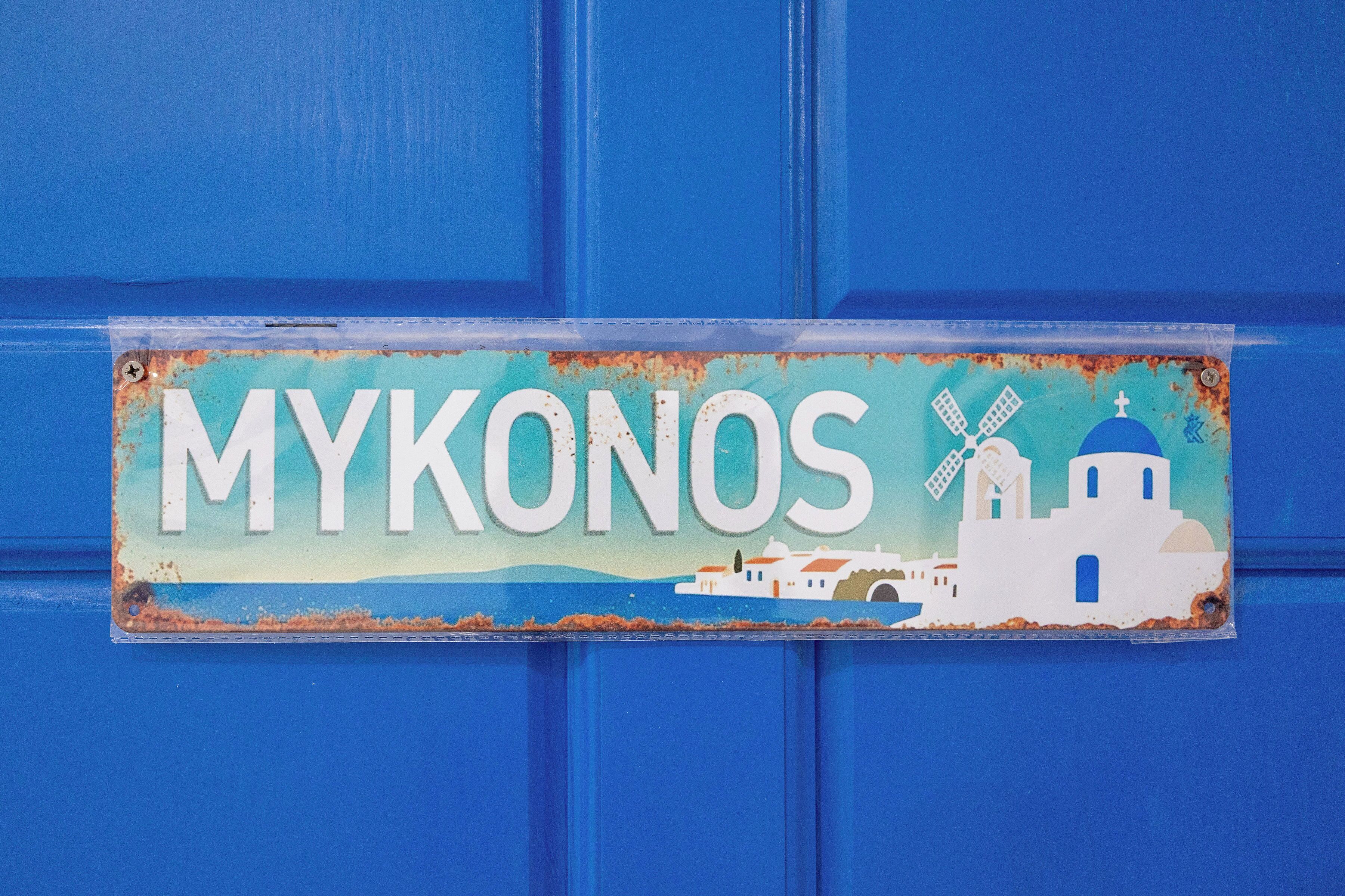 Een bord met de tekst 'MYKONOS' en een afbeelding van een wit gebouw met een blauwe koepel, windmolens en een blauwe achtergrond.