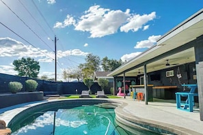 A swimming pool with a curved edge, a covered patio area with colorful stools, and a building with a flat roof. - Scottsdale Hangout | Pool + Putting Green + Games (Scottsdale)