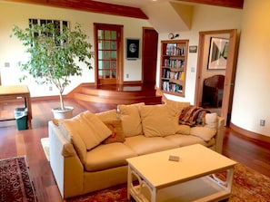 A living room with a beige sofa, a coffee table, a bookshelf, and a potted plant. - Very unique 3-bedroom house on wooded lot near town with great view and privacy. (Homer)