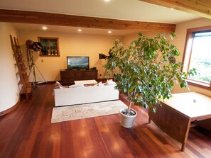 A room with wooden flooring, a large potted plant, a white sofa, a wooden desk, and a television on the wall. - Very unique 3-bedroom house on wooded lot near town with great view and privacy. (Homer)