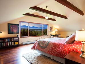 A bedroom with a large window, a bed with a red floral bedspread, a bookshelf, and a view of mountains. - Very unique 3-bedroom house on wooded lot near town with great view and privacy. (Homer)