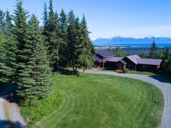 A residential property with a well-maintained lawn, a driveway, and a house with a dark roof. - Very unique 3-bedroom house on wooded lot near town with great view and privacy. (Homer)