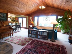 A cozy living room with a wooden ceiling, a piano, a bookshelf, and a large window with a view. - Very unique 3-bedroom house on wooded lot near town with great view and privacy. (Homer)