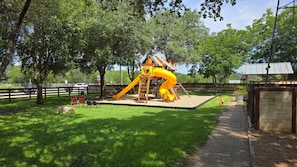 A playground with a slide and climbing structure, surrounded by trees and a fenced area. - Sweetwater Gruene Cottages (New Braunfels)