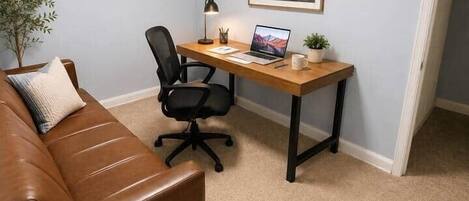 Een kamer met een bruine leren bank, een houten bureau met een laptop, een stoel en een lijst met een afbeelding aan de muur.