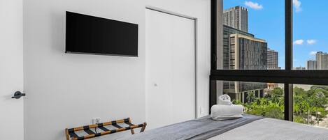 Une chambre à coucher moderne, avec un lit, un téléviseur fixé au mur et une vue sur le paysage urbain grâce à de grandes fenêtres.