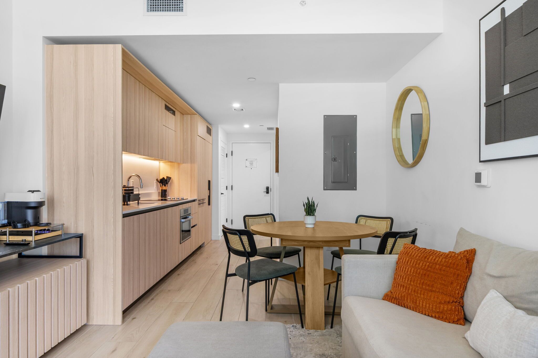 A modern kitchen with wooden cabinets, a dining table with chairs, and a sofa with an orange pillow.