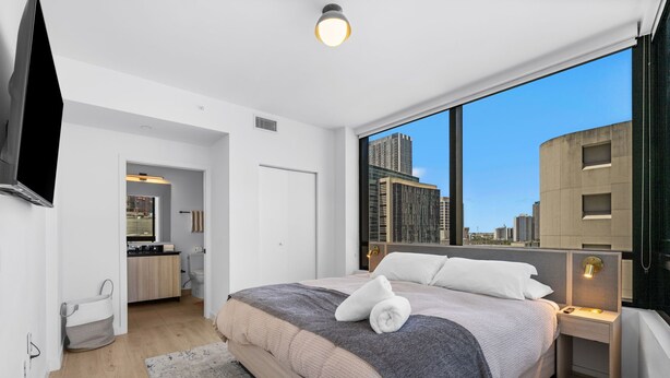 Un dormitorio moderno con una cama grande, vistas de la ciudad, un baño y un televisor montado en la pared.