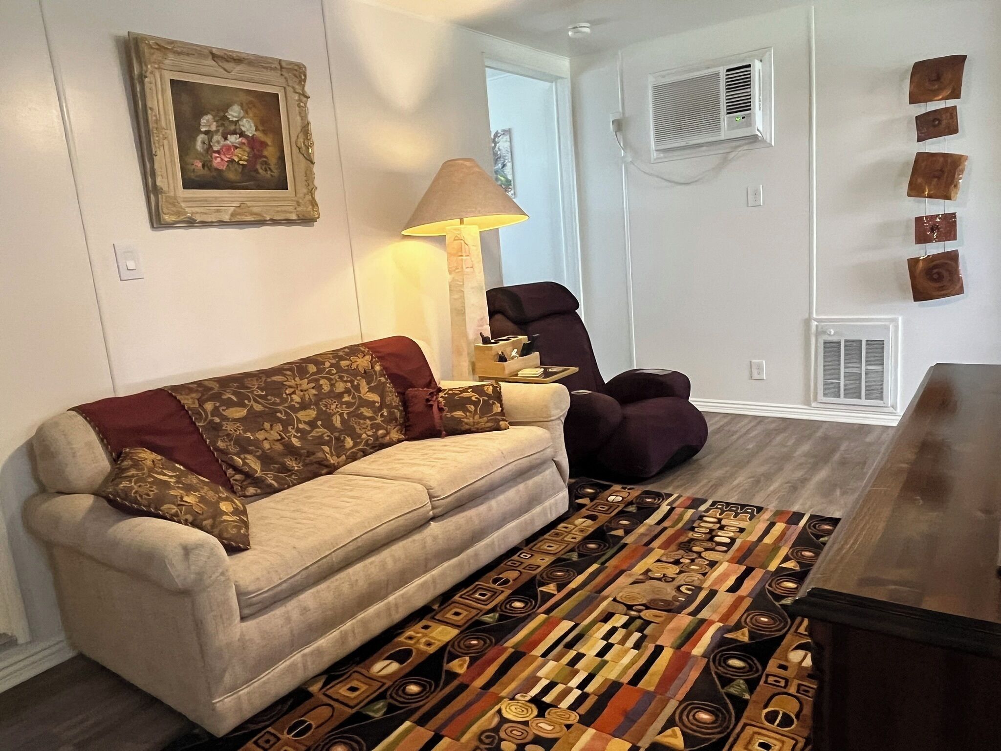 A living room with a sofa, a chair, a patterned rug, a lamp, and a wall-mounted air conditioning unit.