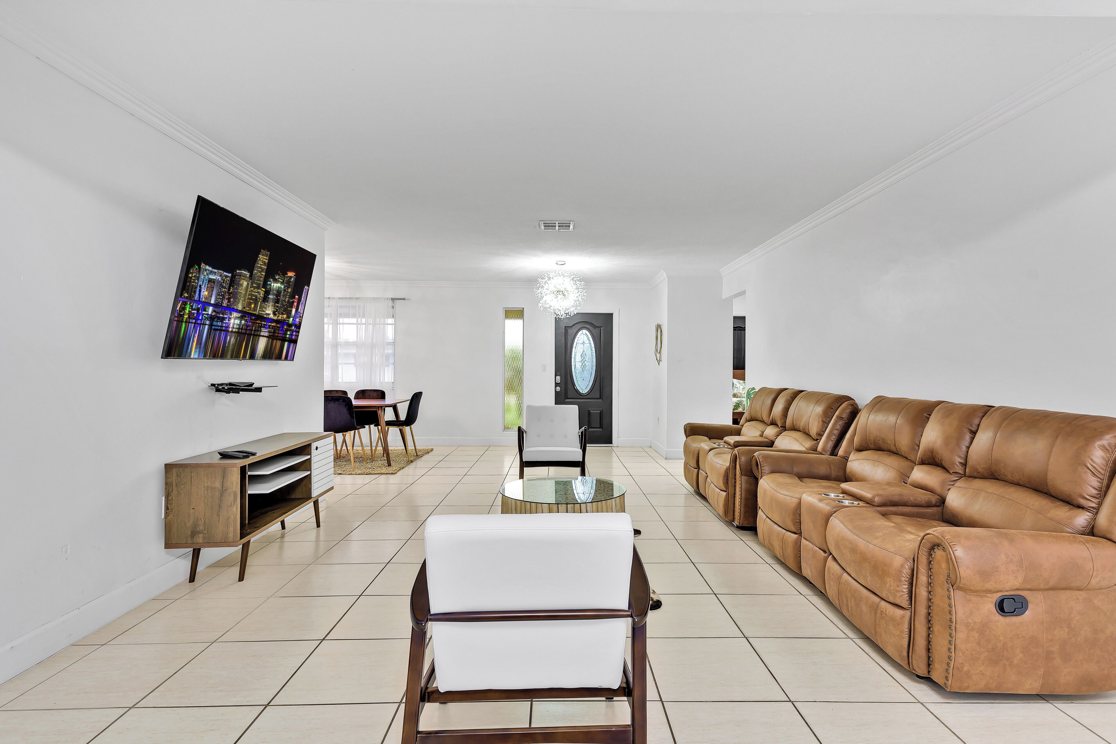 Una sala de estar amplia con un sofá grande de cuero, un sillón blanco, una mesa de centro con superficie de vidrio y un televisor de pantalla plana montado en la pared.