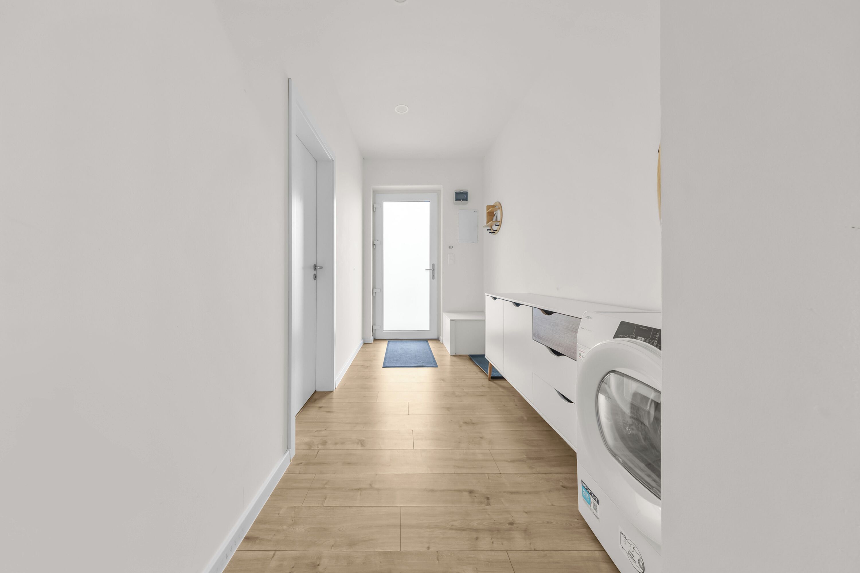 A laundry room with a wooden floor, white walls, and a door leading to another room.