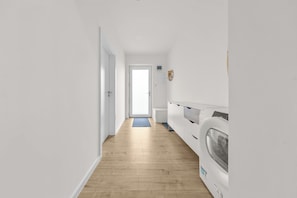 Una sala de lavandería con piso de madera, paredes blancas y una puerta que da a otra habitación.