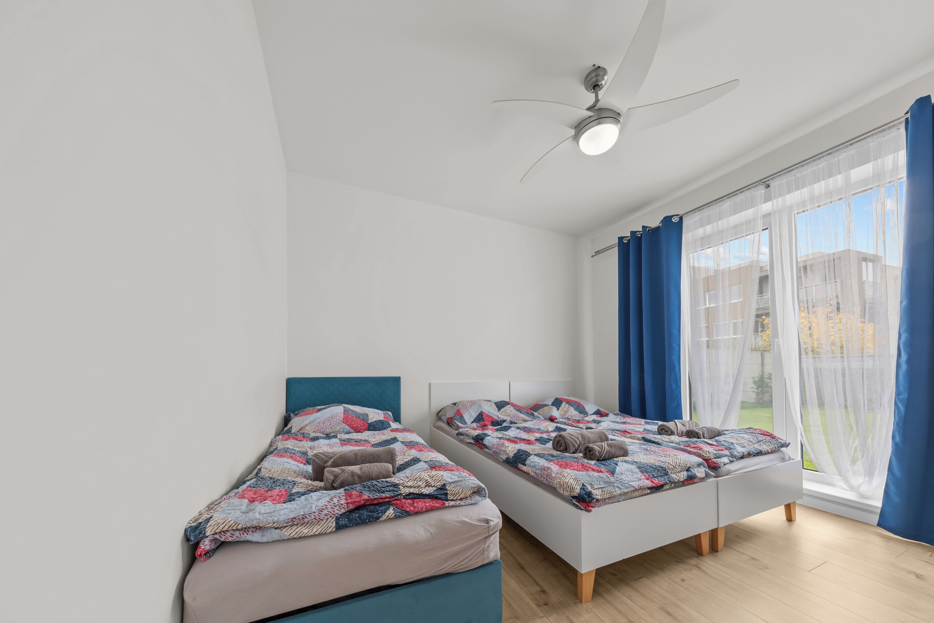 A modern bedroom with two single beds, a ceiling fan, and a large window with blue curtains.