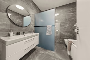 A modern bathroom with a white vanity, a round mirror, a blue shower enclosure, and a toilet.