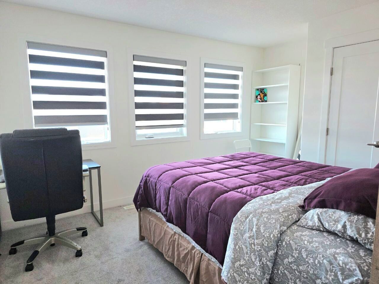 Un dormitorio con una cama, un escritorio y una silla. Hay tres ventanas con persianas.