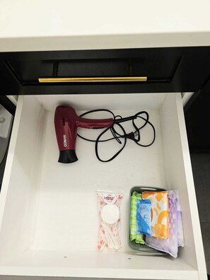 Un tiroir contenant un sèche-cheveux rouge, un câble noir, une bouteille blanche et divers articles en tissu de couleurs vives.