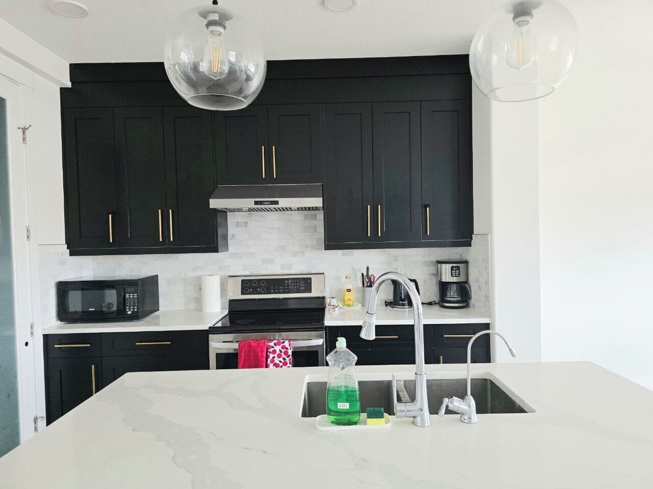 A modern kitchen with dark cabinetry, a white countertop, a built-in microwave, a stainless steel range hood, and a double sink.