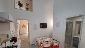 Een kleine keuken met een fornuis, een eettafel met stoelen, een aan de muur bevestigde televisie en een douchehoek.