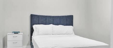 A single bed with a headboard, a bedside table with drawers, and a window with blinds.