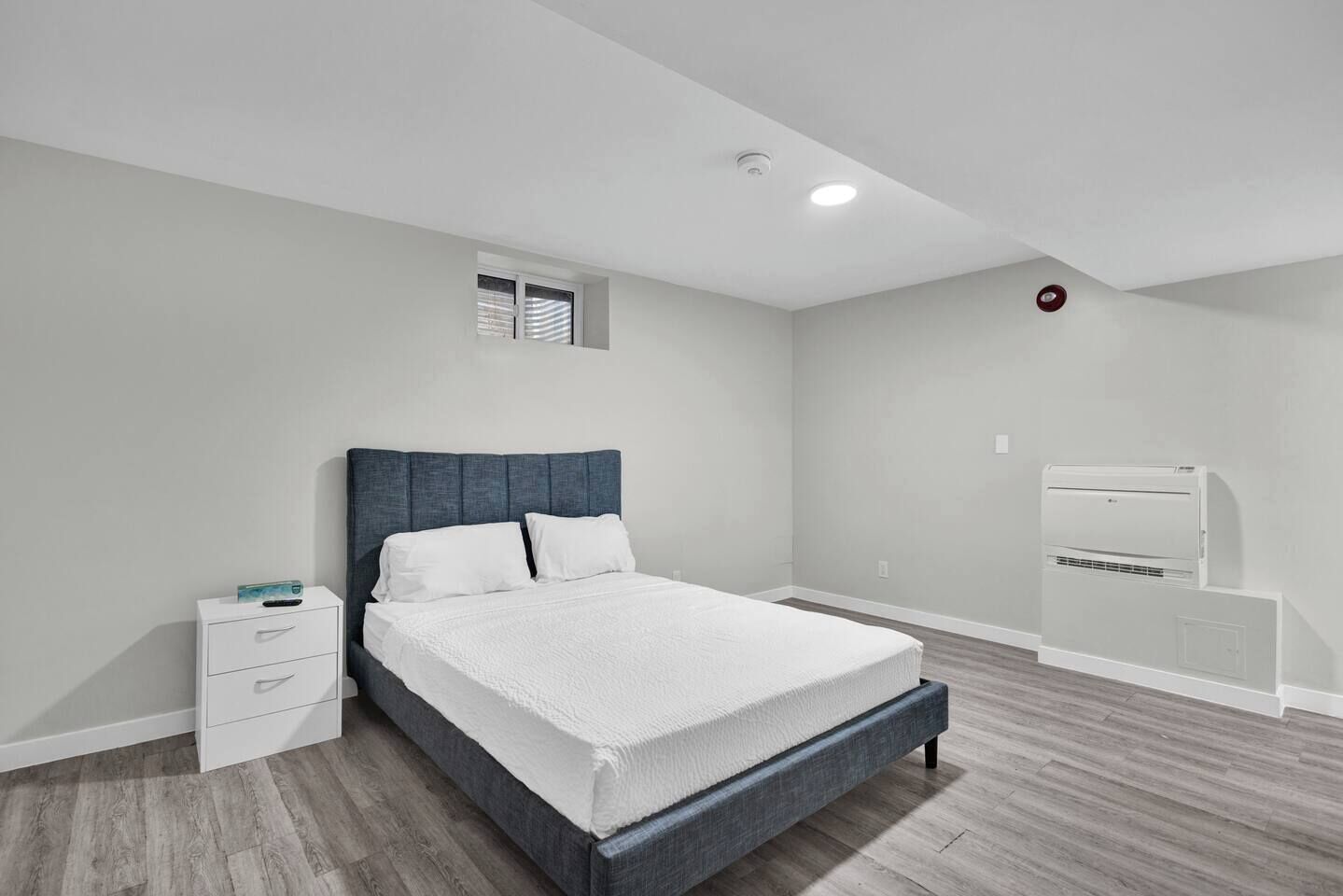 Un dormitorio con cama, mesita de noche, ventana y equipo de aire acondicionado.