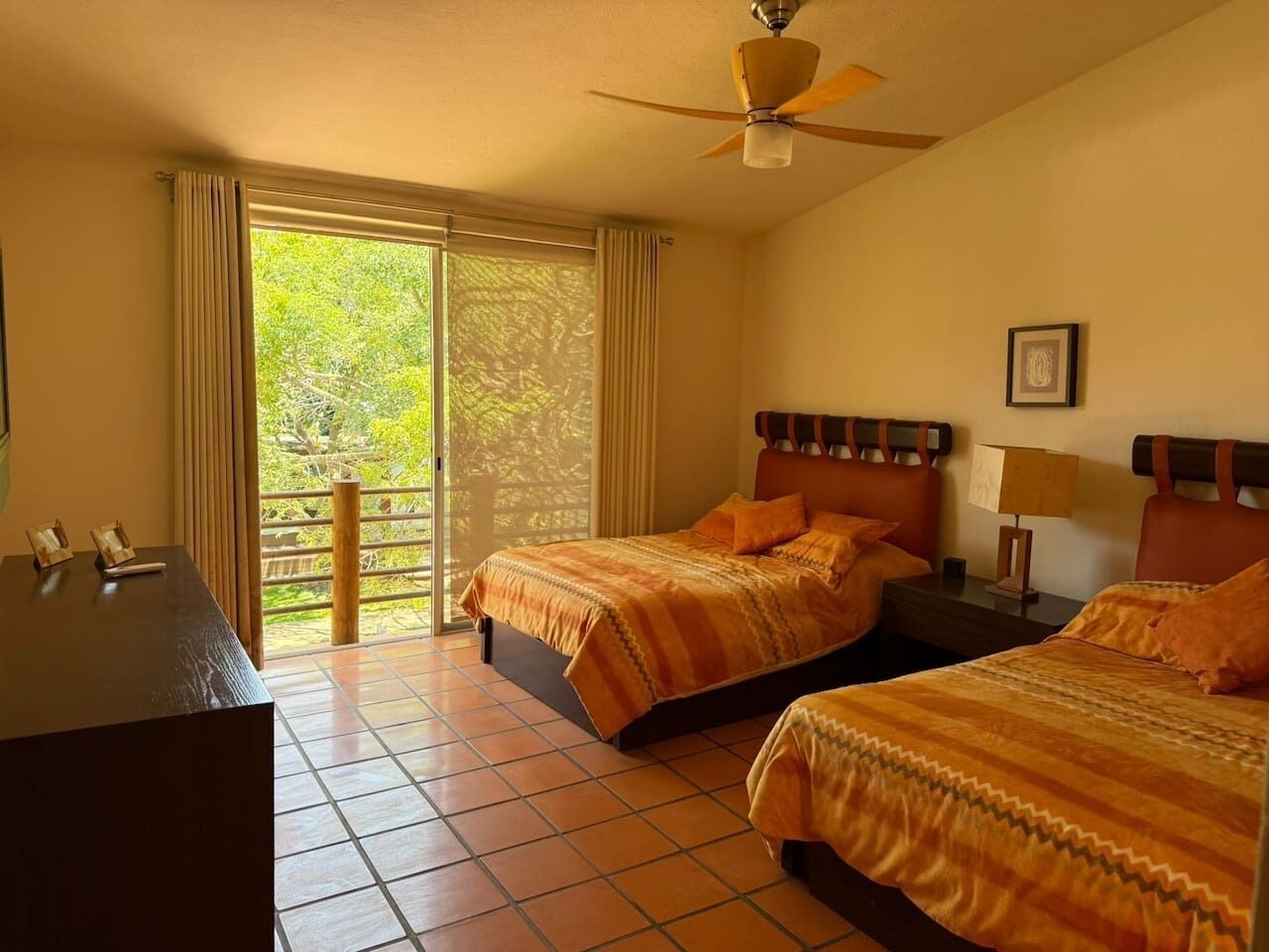 A hotel room with two beds, a desk, a ceiling fan, and a view of greenery through the window.