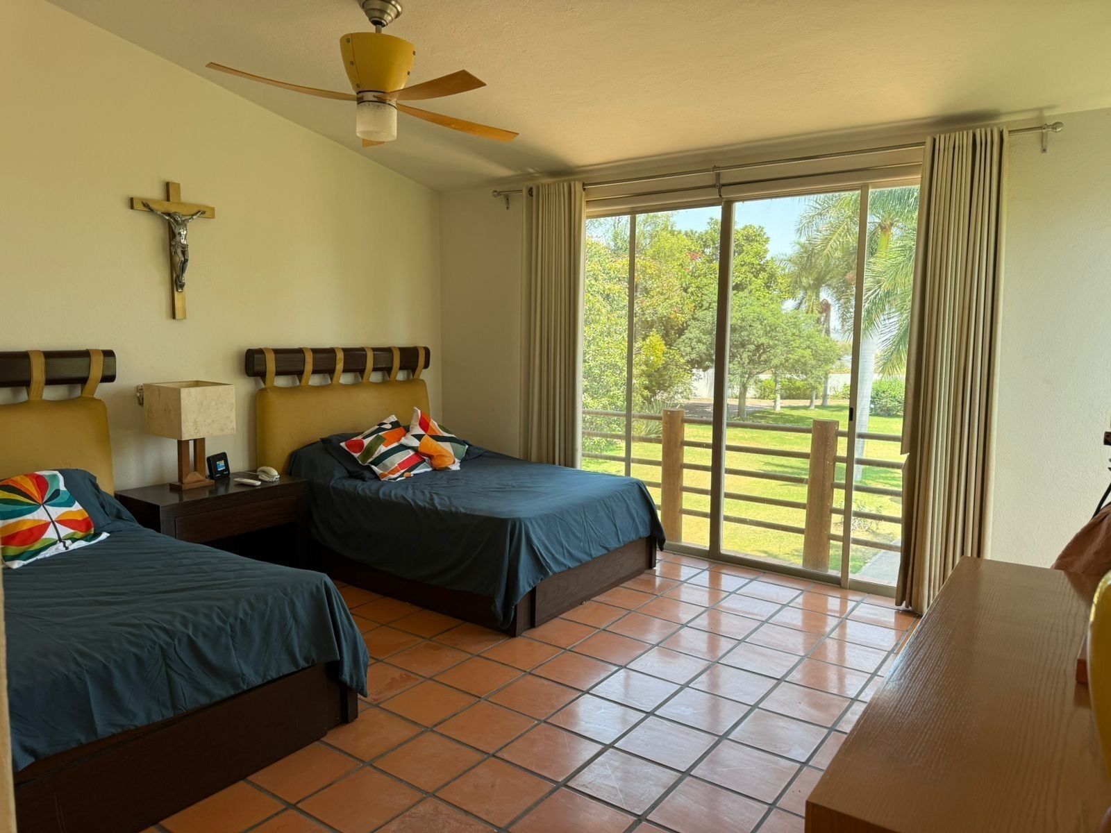 A hotel room with two beds, a ceiling fan, a cross on the wall, and a view of a garden through the window.