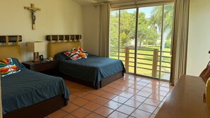Une chambre d’hôtel avec deux lits, un ventilateur de plafond, une croix accrochée au mur et une vue sur un jardin par la fenêtre.