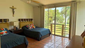 A hotel room with two beds, a ceiling fan, a cross on the wall, and a view of a garden through the window.