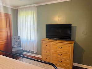 A room with a bed, a wooden dresser with drawers, a television on top, a chair, and a window with curtains.