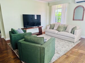 Ein Wohnzimmer mit Flachbildfernseher, grünen Sessel, beiger Couch und einem großen Fenster mit Vorhängen.