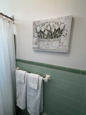 A bathroom with a green tiled wall, a white towel rack with two towels, and a framed black and white floral painting above the towel rack.