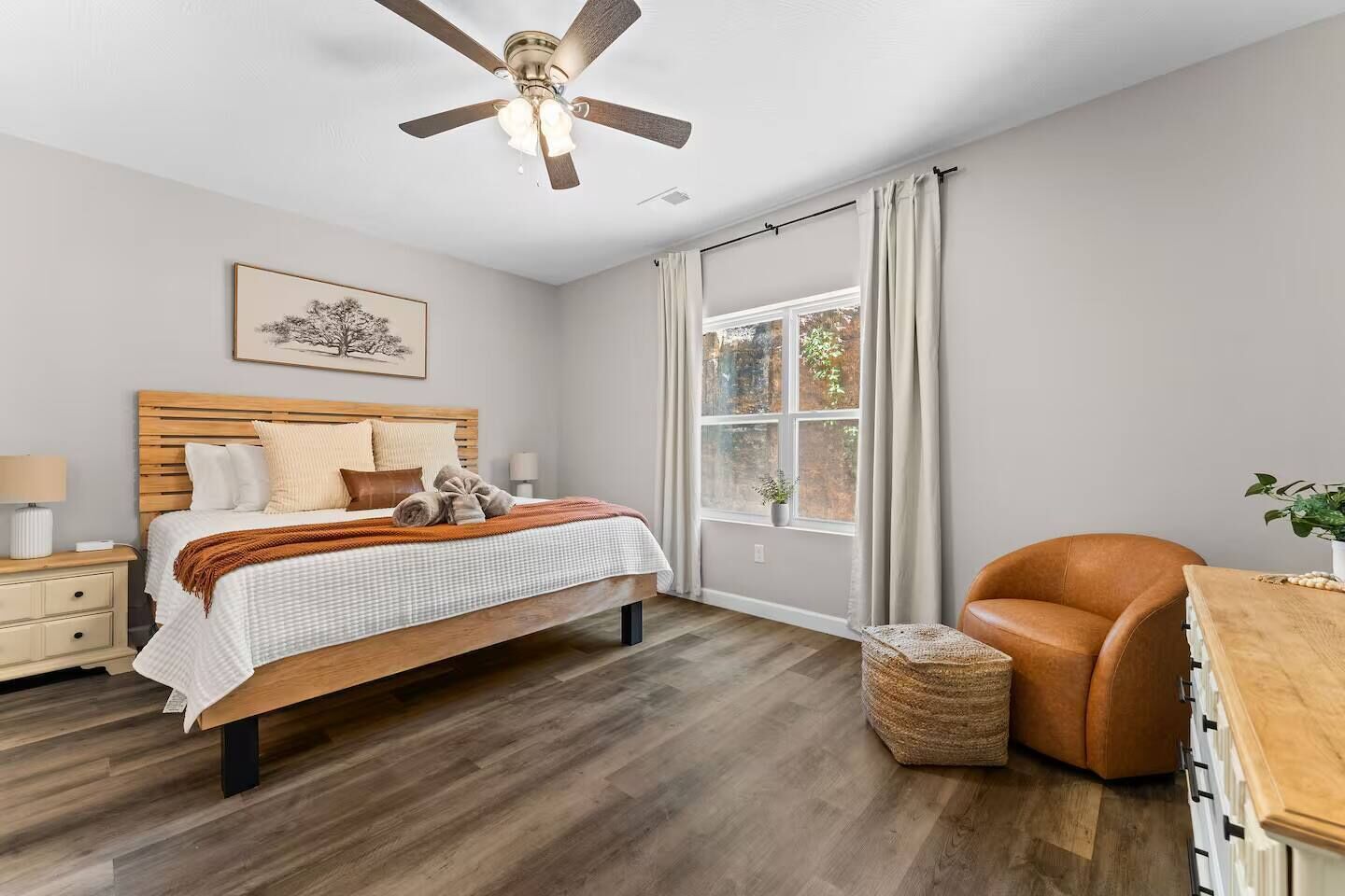 A bedroom with a bed, bedside table, lamp, ceiling fan, window with curtains, and a chair.