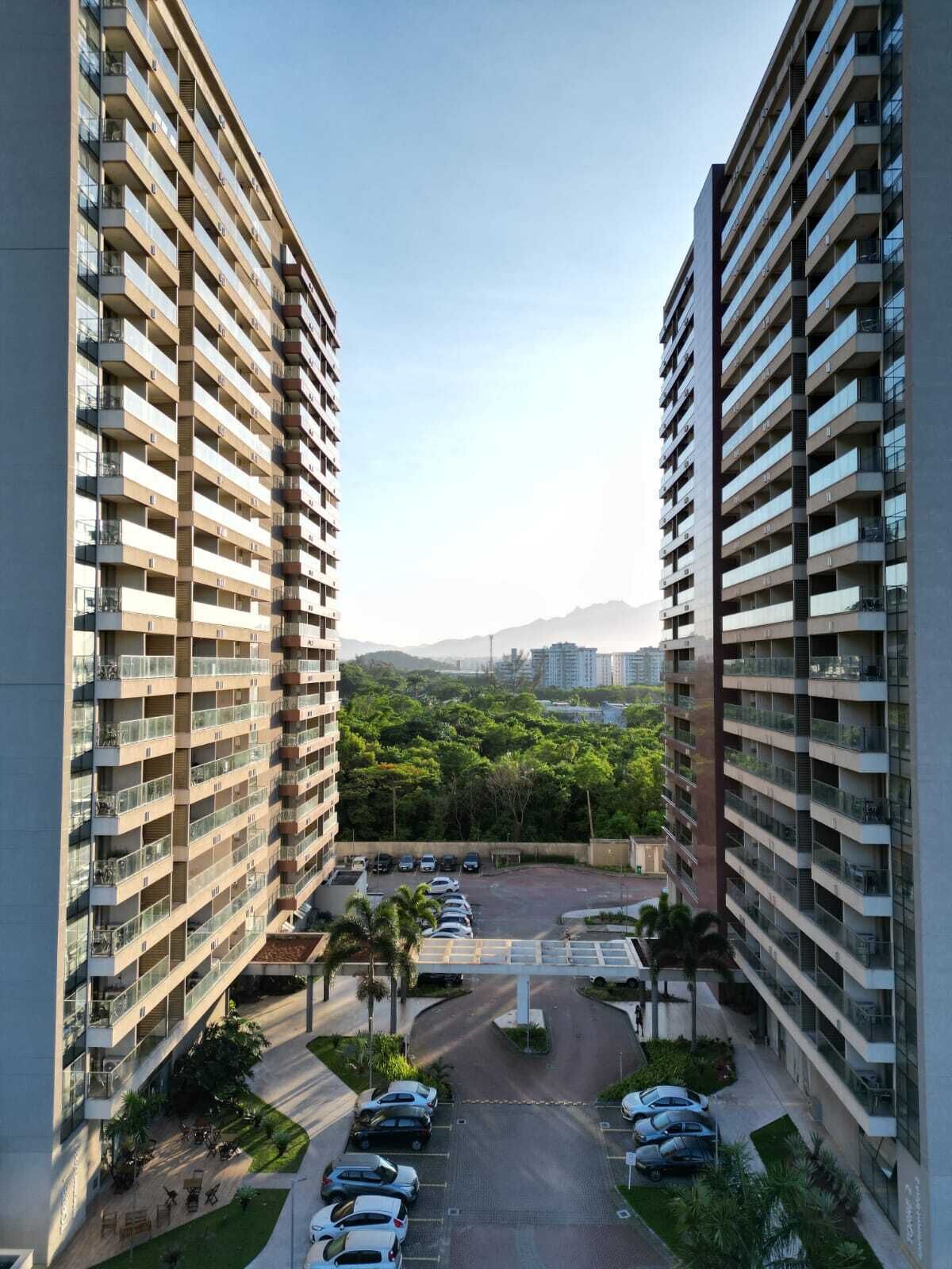 Un edificio residencial alto con varios balcones, un área de estacionamiento y vistas a un paisaje verde con montañas al fondo.