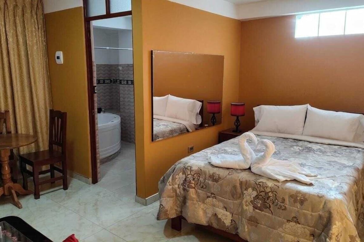 Habitación de hotel con cama, una silla, una mesa, un espejo y un baño visible a trav és de una puerta abierta.