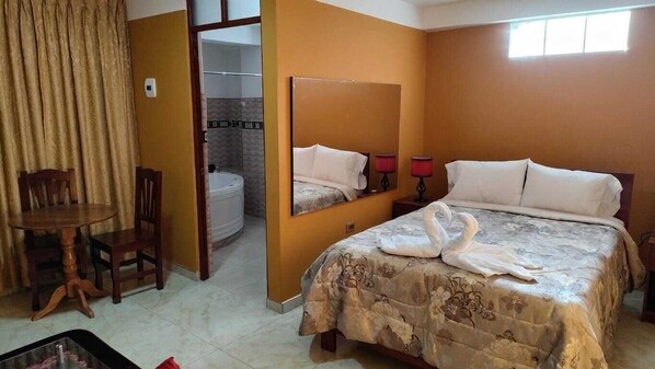 Habitación de hotel con cama, una silla, una mesa, un espejo y un baño visible a través de una puerta abierta.