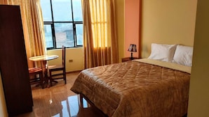 A hotel room with a bed, a small round table, a chair, a lamp, and a view through a window.