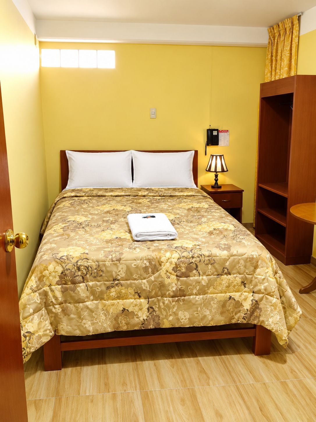 A bed with a floral bedspread, a bedside lamp, a wooden wardrobe, and a door with a golden handle.
