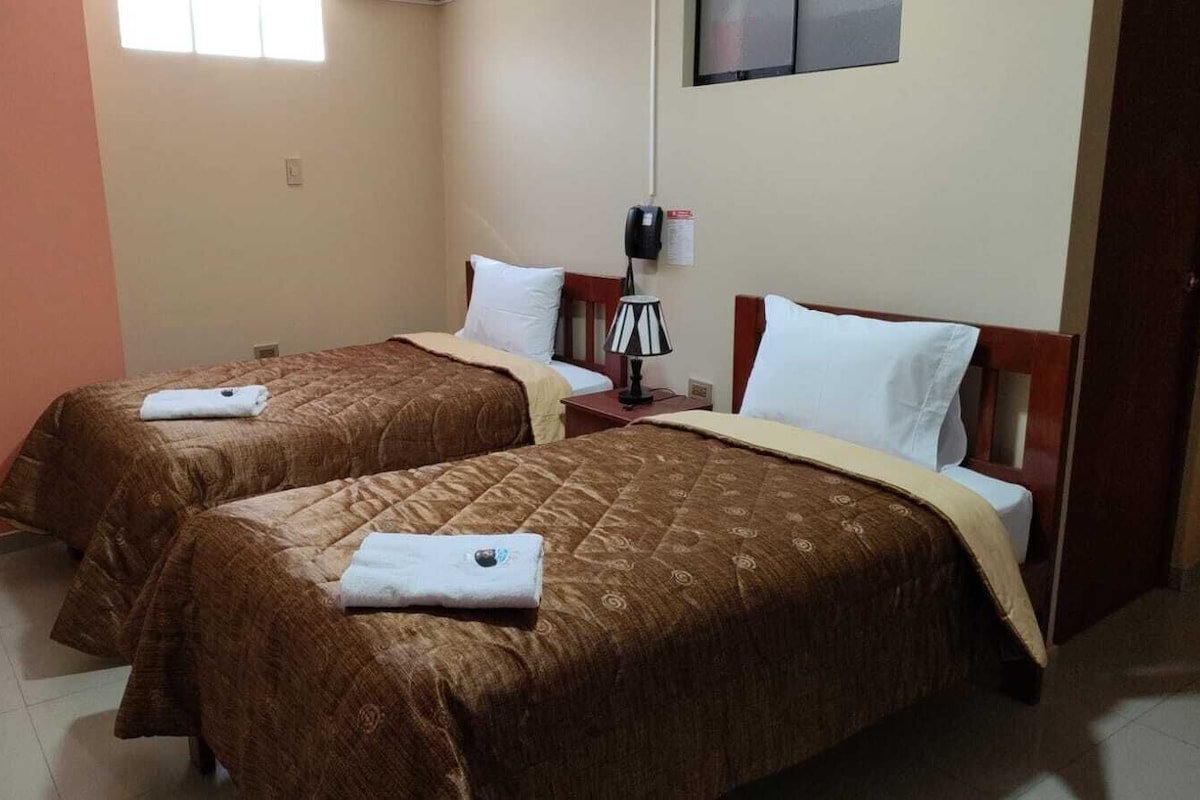 A hotel room with two beds, each with a brown quilt and white pillows, a bedside table with a lamp, and a wall-mounted phone.