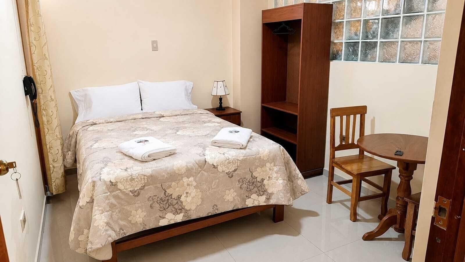 Un dormitorio con cama, mesita de noche, armario de madera, una silla y una pequeña mesa.