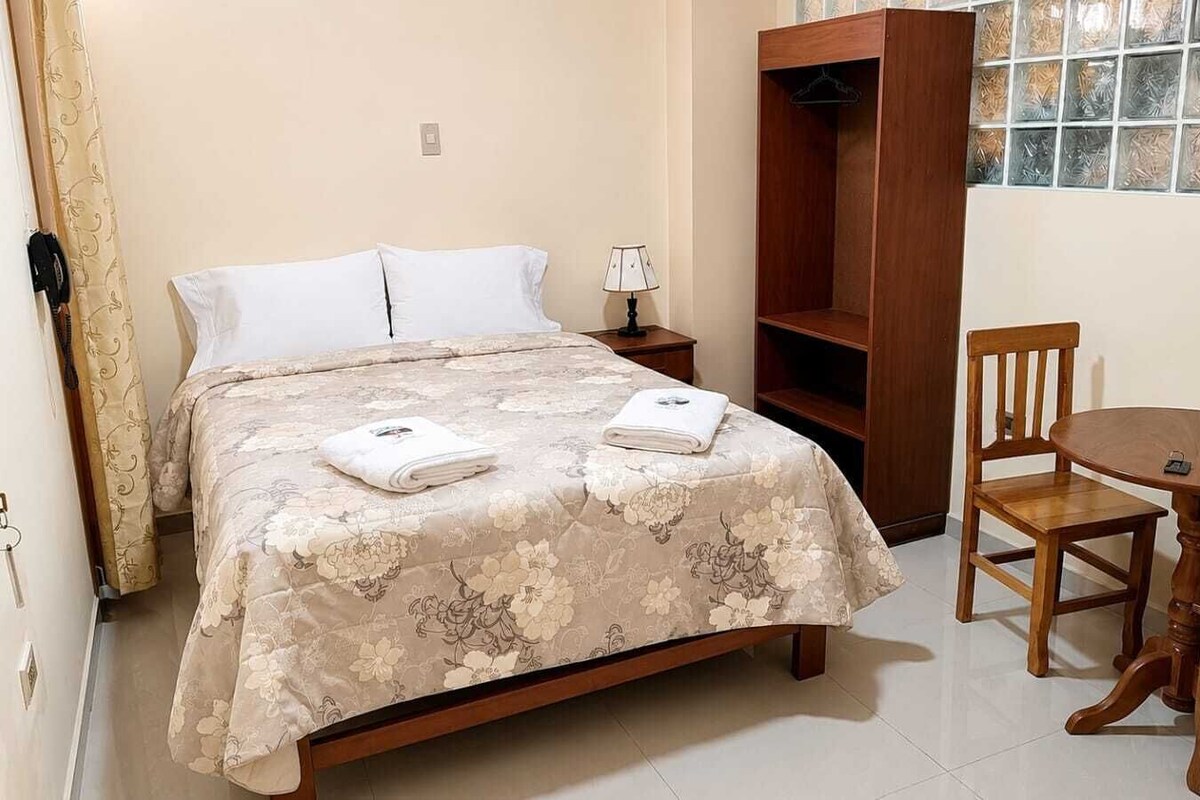 Un dormitorio con cama, mesita de noche, armario de madera, una silla y una pequeña mesa.