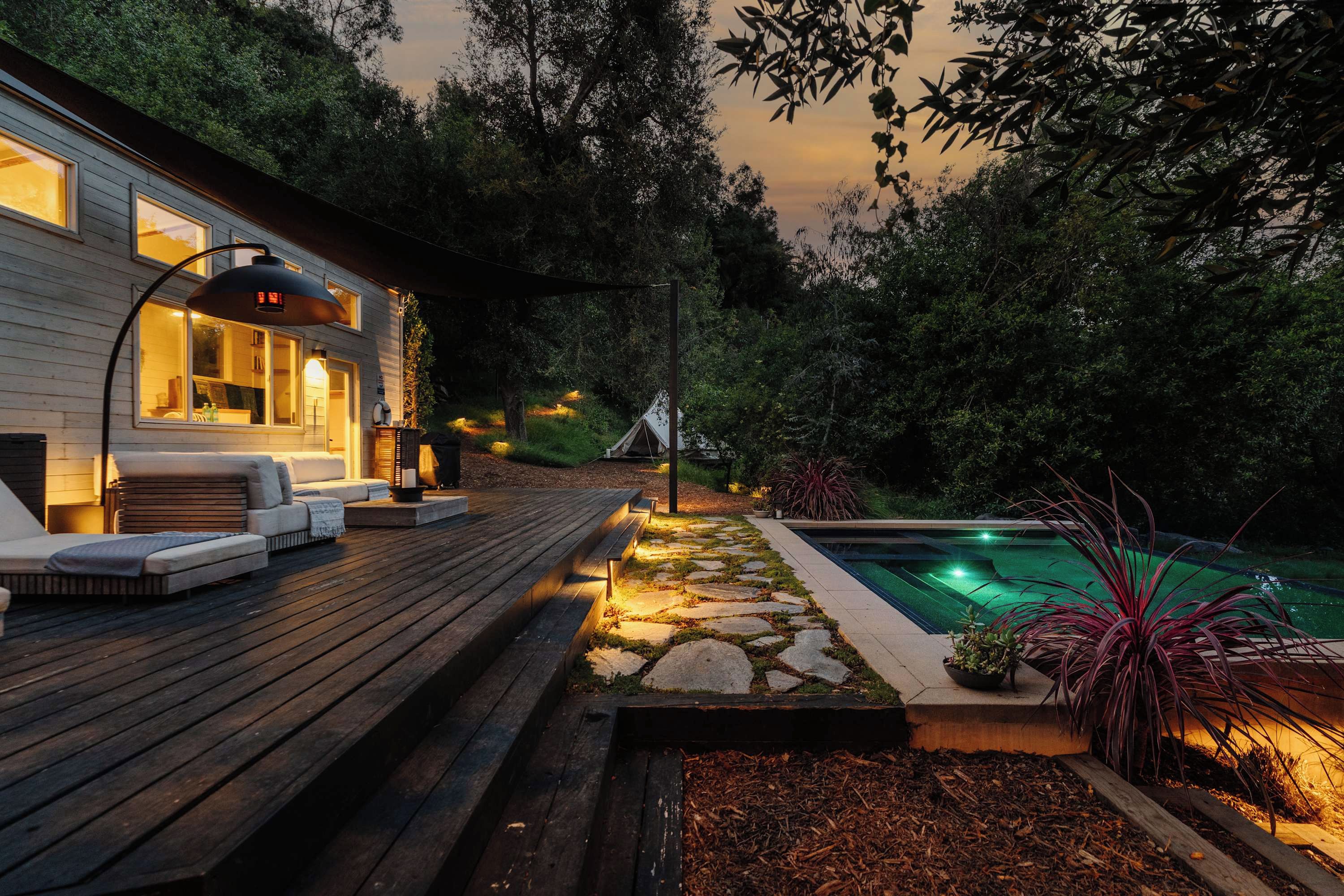 A wooden deck with outdoor seating, a swimming pool, and a house with illuminated windows.