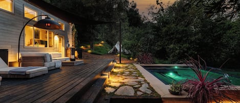 A wooden deck with outdoor seating, a swimming pool, and a house with illuminated windows.