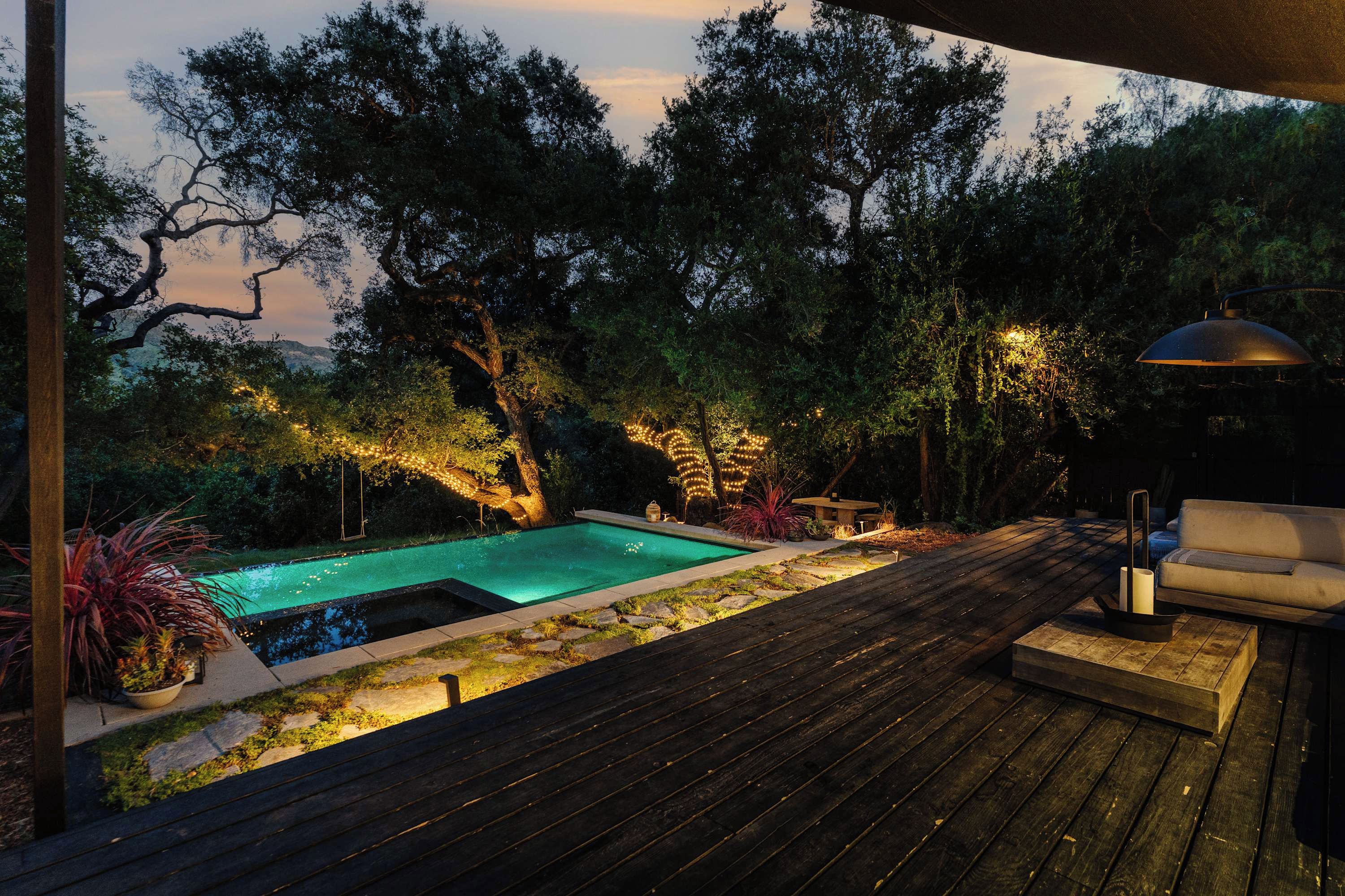 Una terrassa de fusta amb piscina, envoltada d'arbres i il·luminada per llums de cadena.
