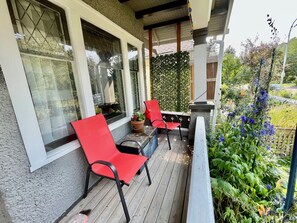 Eine Holzterrasse mit zwei roten Stühlen und einem Tisch, ein Fenster mit weißem Rahmen und eine grüne, blätterreiche Wand.
