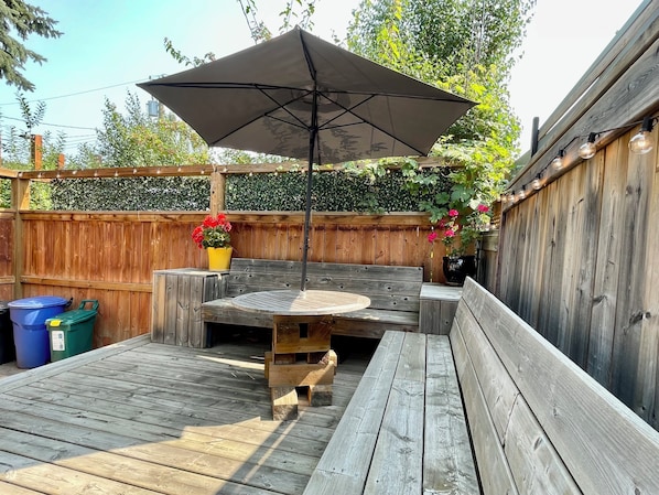 Eine Holzterrasse mit rundem Tisch und Bänken, ein Pergola mit Lichterketten und ein Holzzaun mit Grünpflanzen.