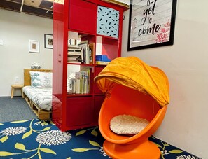 Ein Zimmer mit einem roten Bücherregal, einem kleinen Holztisch, einem Bett mit gemustertem Überwurf und einem orangefarbenen Sessel mit Kissen.